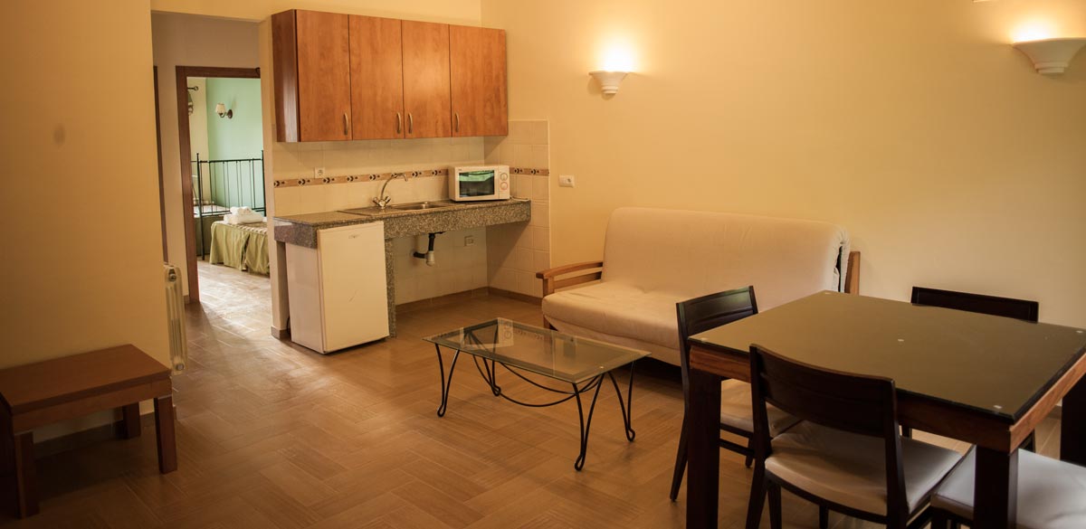 Salón con cocina completa de uno de los apartamentos del Complejo Rural Puerto Mágina. Una mesa y sillas para 4 personas. Cocina con frigorífico, microondas, fregadero y muebles con utensilios. Muestra radiador de calefacción, sofá y mesilla de café de cristal.