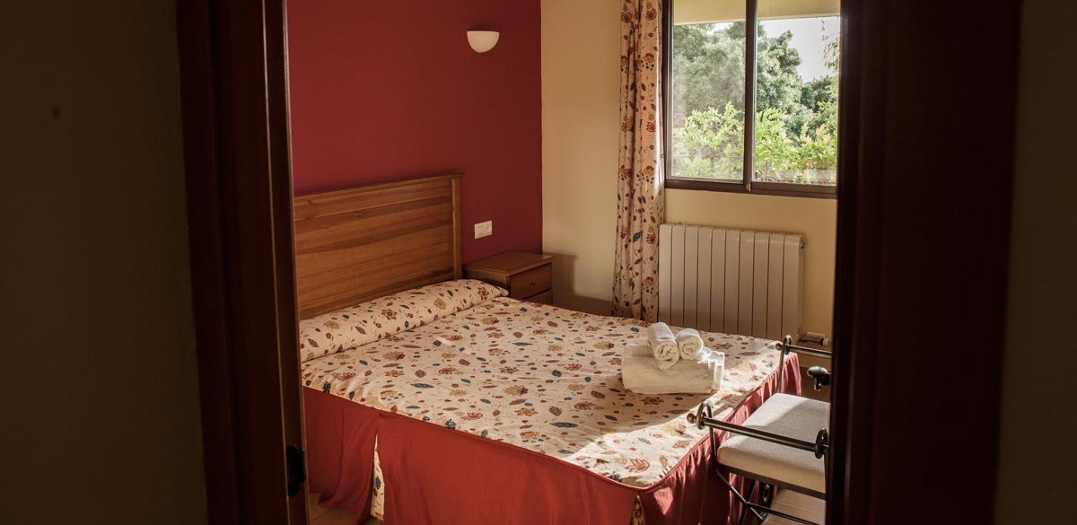 Una habitación de uno de los apartamentos del Complejo Rural Puerto Mágina. Con ventana con vistas a Sierra Mágina. Muebles de madera noble. Colcha y cortinas estampadas con motivos florales destacando los colores rojizos.