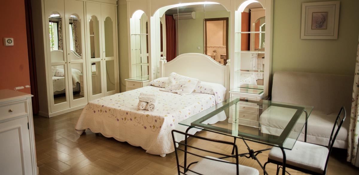 Fotografía de uno de los dormitorios del Complejo Rural Puerto Mágina. Es de mobiliario blanco y tiene muchos espejos. Se muestra, también, un pequeño sofá y una mesa de cristal cuadrada con dos sillas de forja con cojines.