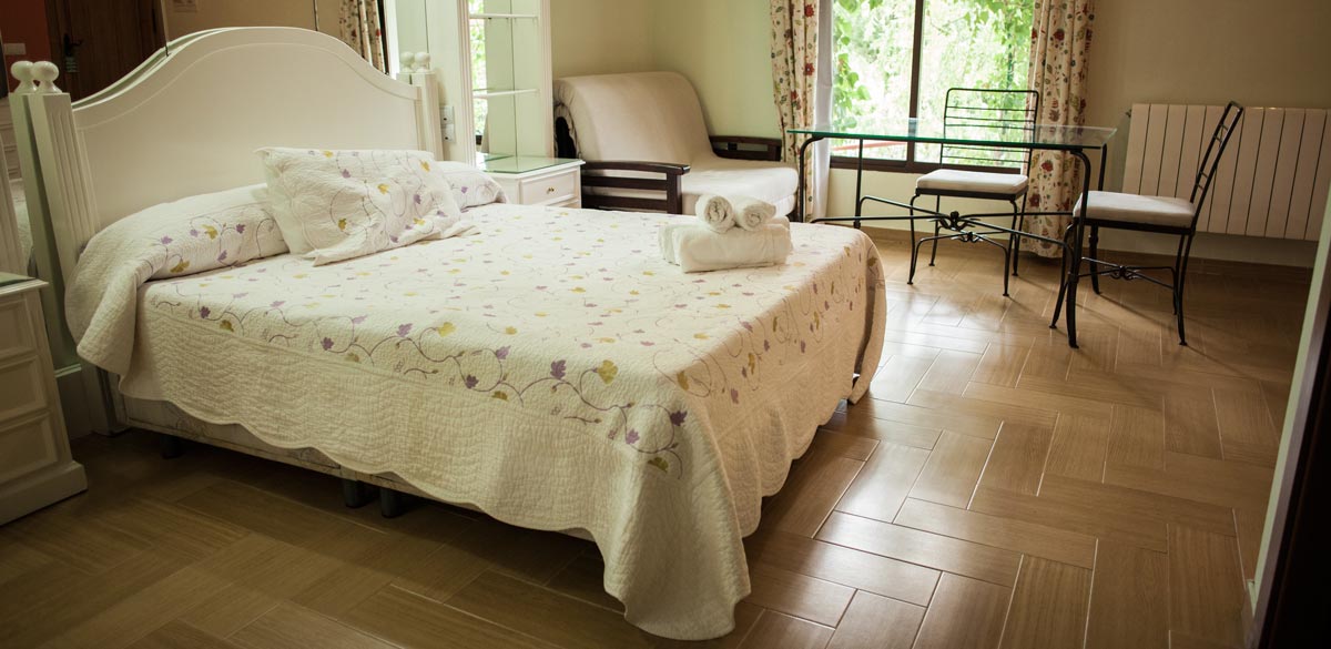 Destalles de una de las habitaciones del Complejo Rural Puerto Mágina. Muebles, blancos. Una cama de matrimonio, un pequeño sofá y dos sillas. Al fondo un gran ventanal con vista a Sierra Mágina.