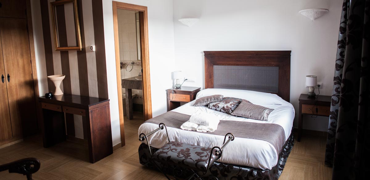 Habitación de madera con tonos marrones. Una cama de matrimonio y varios muebles auxiliares. Se muestra la puerta de acceso al aseo de esta habitación del Complejo Rural Puerto Mágina.