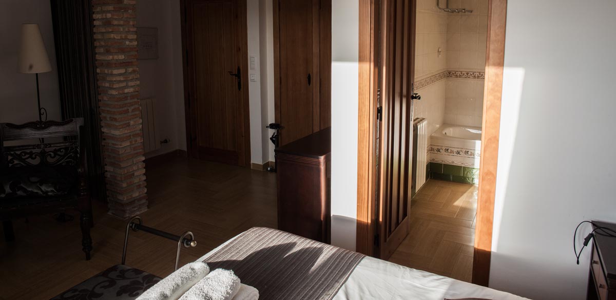 Fotografía desde la cama de matrimonio donde se muestran accesos a la salida y un aseo de una de las habitaciones del Complejo Rural Puerto Mágina. También se muestran las puertas de un armario empotrado.