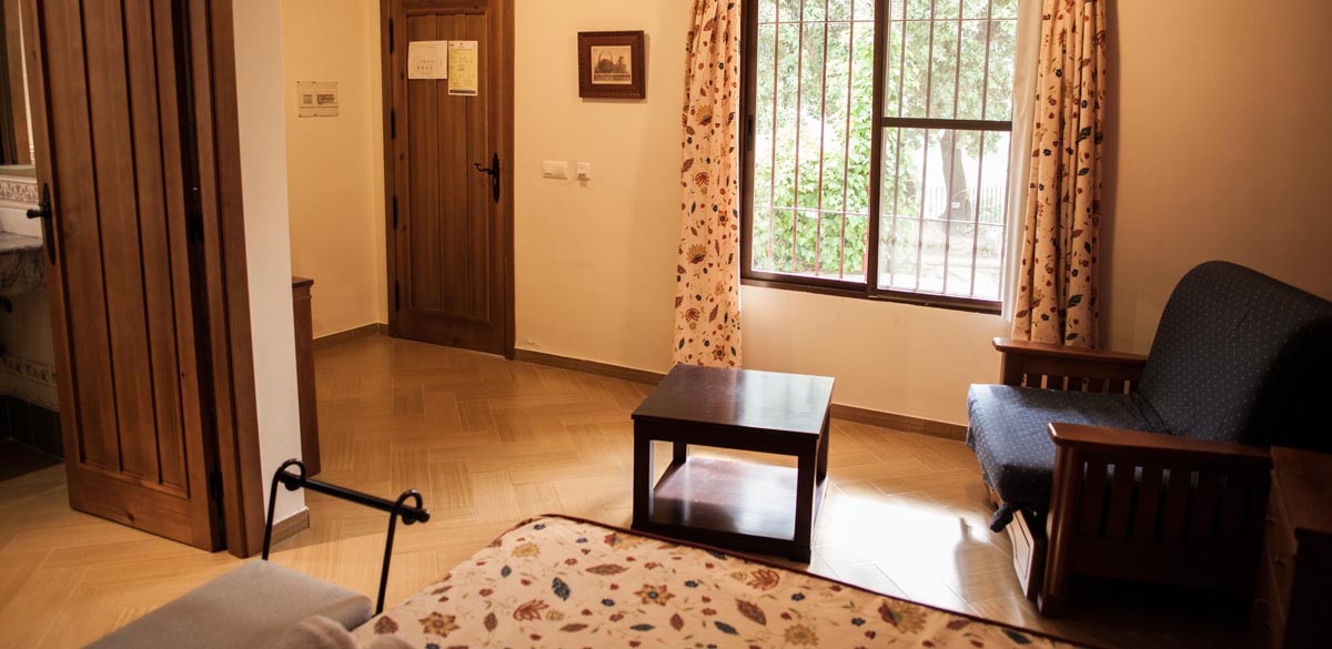 Pequeño hall de una de las habitaciones del Complejo Rural Puerto Mágina. Un sofá individual junto a una mesita de madera. Se muestra un gran ventanal con vista a los exteriores.