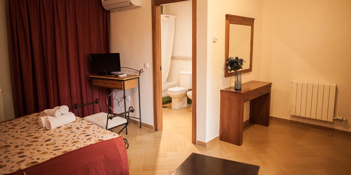 Vista desde un sillón del pequeño hall de una de las habitaciones del Complejo Rural Puerto Mágina. Se ven muebles auxiliares; en uno de ello hay una televisión. Se puede ver la puerta abierta de acceso al aseo.